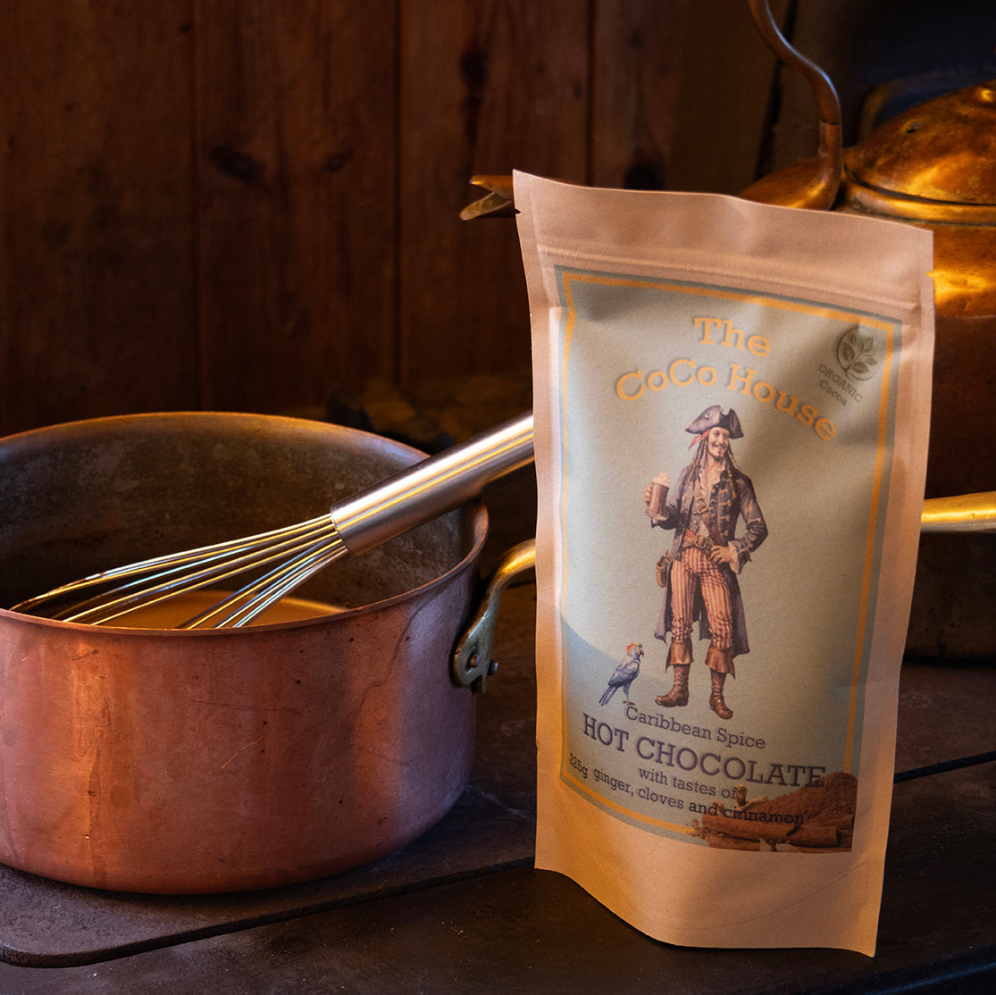 A stand-up pouch of 'The Cocoa House' Hot Chocolate next to a copper pot with a whisk on a rustic wood stove.